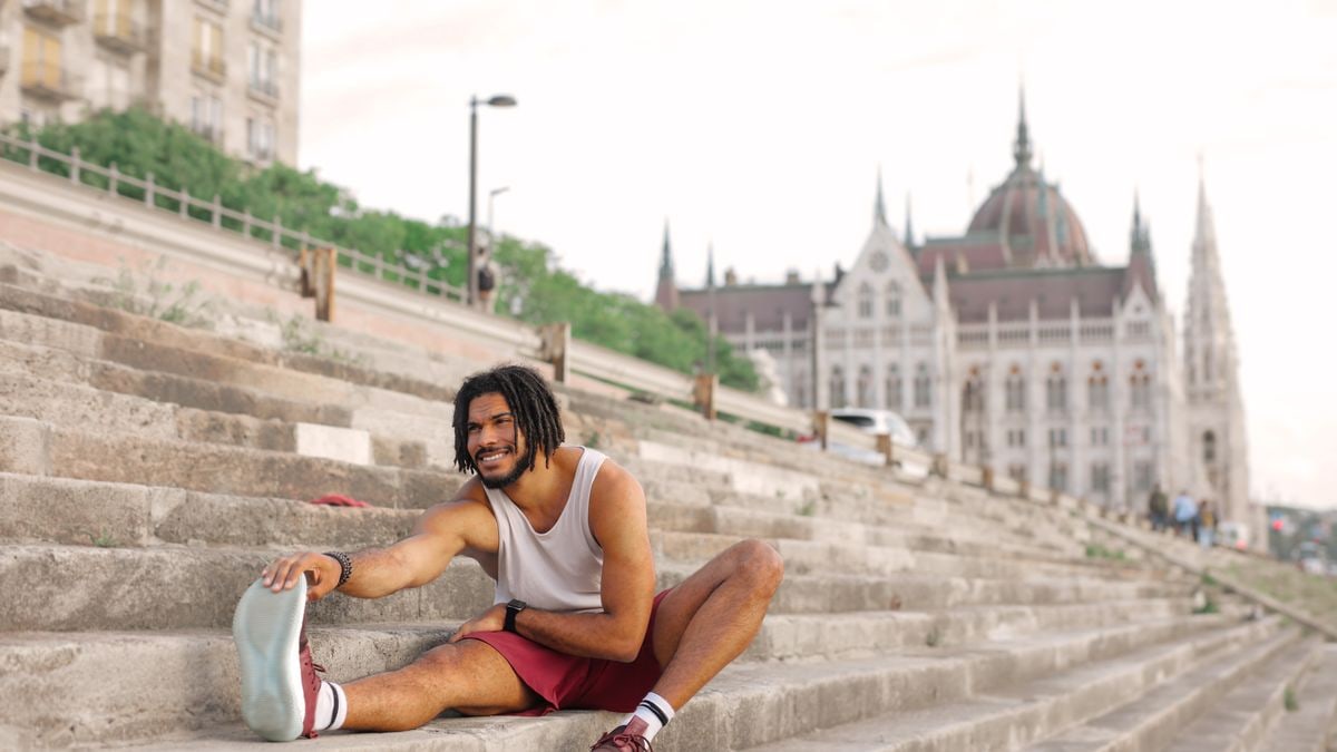 Man with dreadlocks smiling while stretching on city steps near architectural landmark.